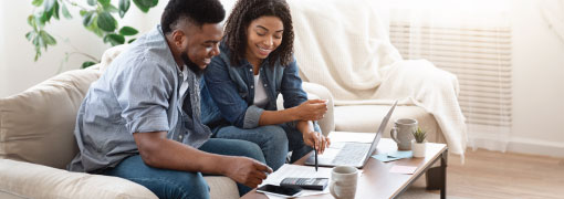 A smiling couple sits on a couch reviewing finances together on the table in a bright living room.