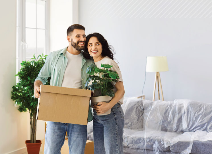 Happy couple moving into their new home holding a moving box and houseplant with furniture