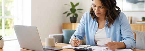 Woman reviewing receipts and writing in a budget notebook beside a laptop and calculator at home.