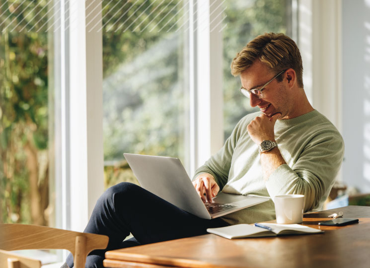 A man sits by a bright window, smiling while reviewing FSB’s CD rates on his laptop, with a notebook and coffee nearby.