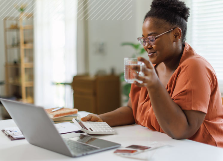 Woman at home reviews bills with a calculator and laptop while holding a glass of water.