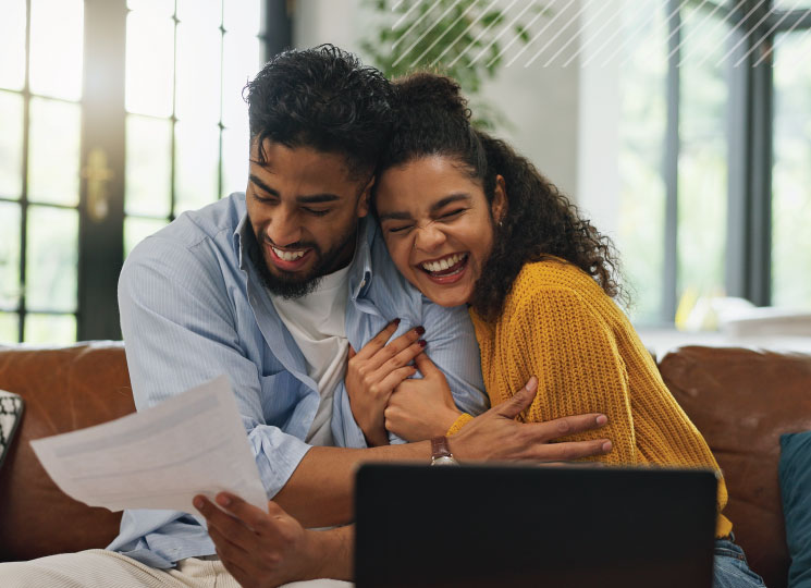 Couple on a couch smiles and hugs while looking at paperwork, with a laptop in the foreground.