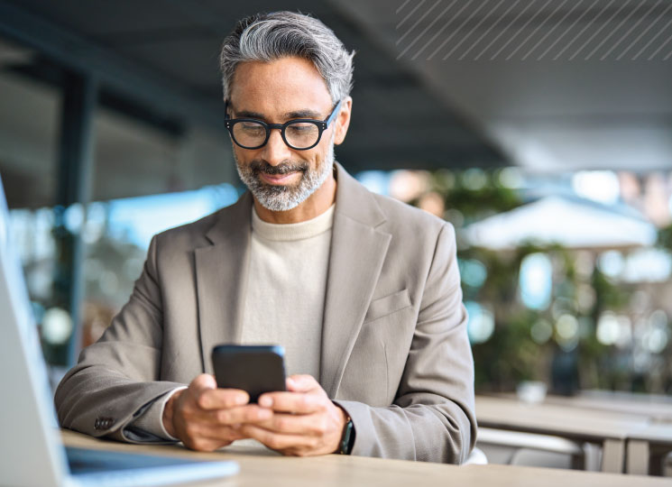 A professional man in glasses looks at his smartphone while sitting at a table with a laptop nearby