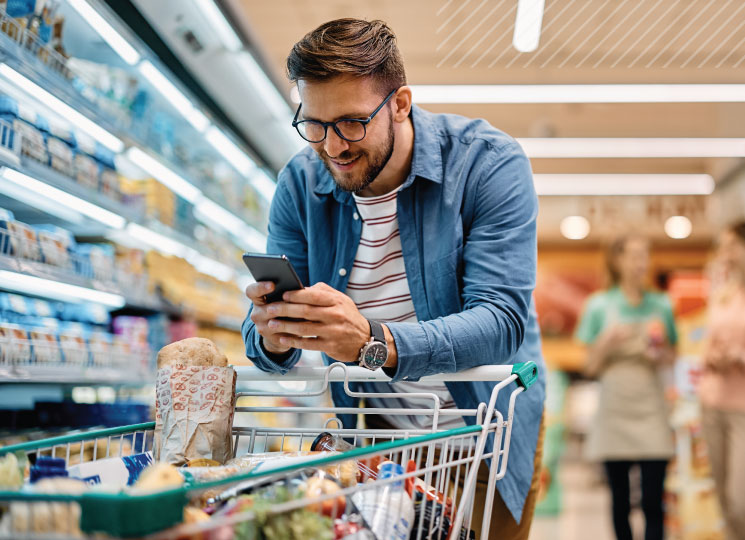 Man in a grocery store smiles while checking his smartphone beside a shopping cart filled with groceries.