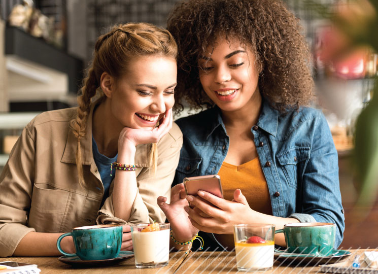 Two friends sit at a café table smiling while looking at a smartphone, with coffee cups and drinks on the table.
